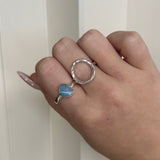 Hand wearing a silver ring with a blue gemstone on a plain background