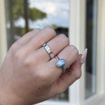 Hand wearing two rings with a blue gemstone in front of a glass door.