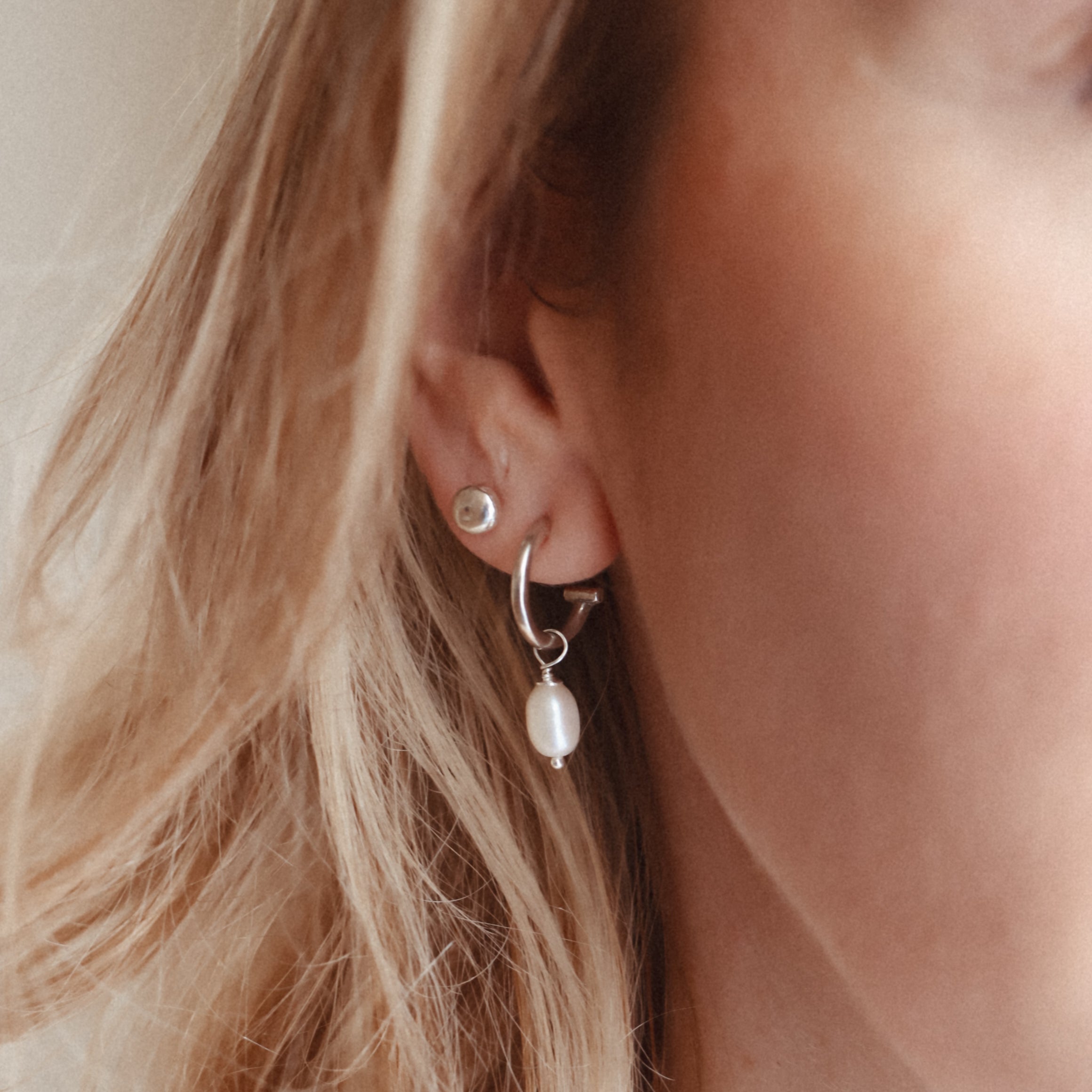 A close-up photo of a woman's ear wearing a silver hoop earring with a pearl charm.