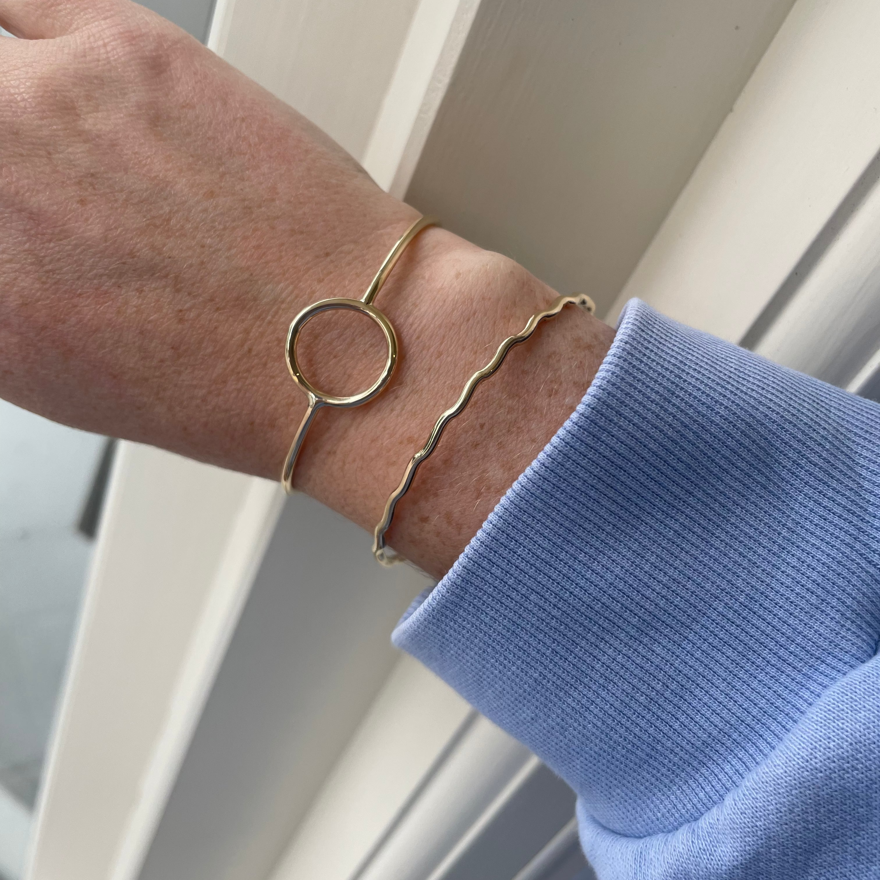 Gold bracelet on a wrist with a blue sleeve against a neutral background