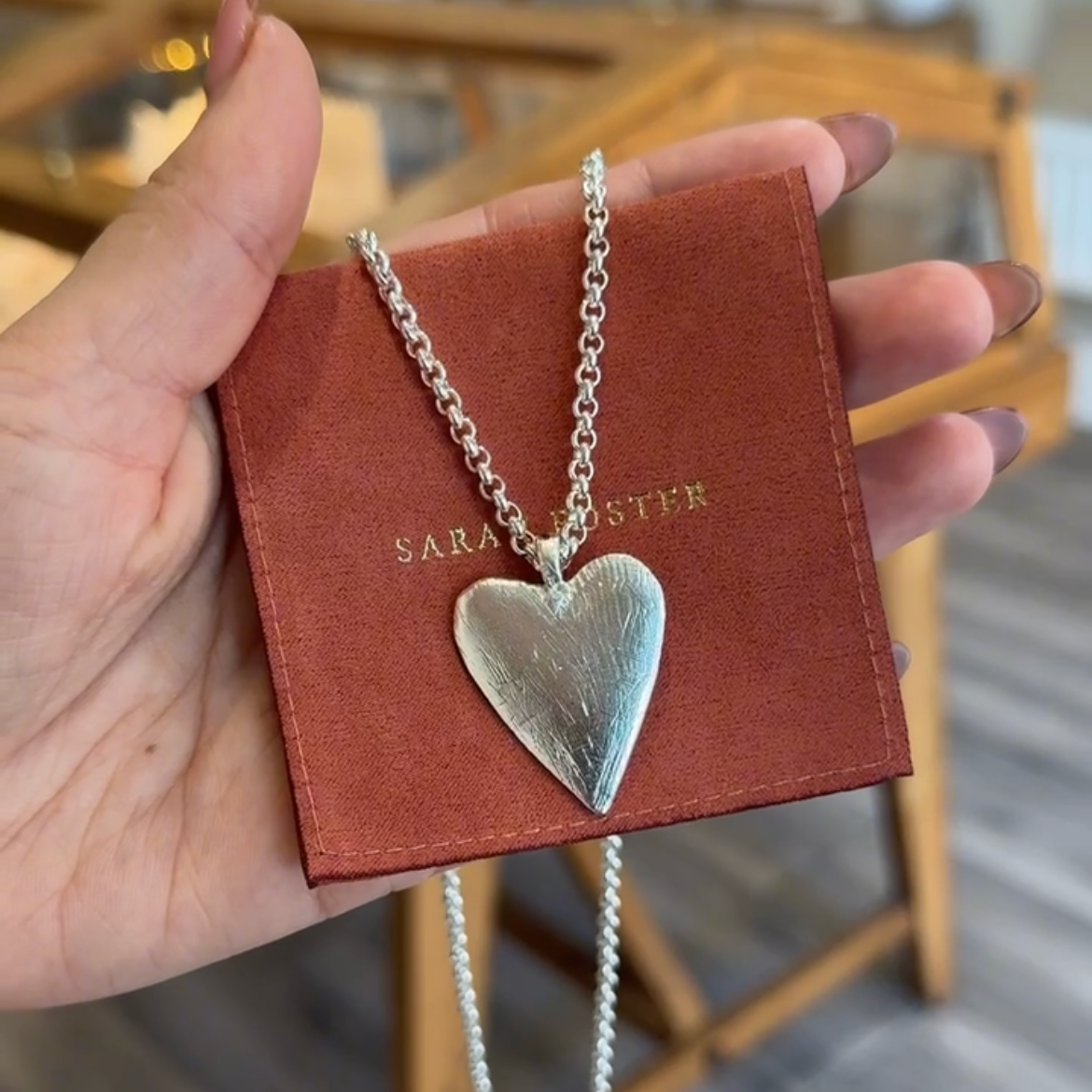 Heart-shaped silver necklace on a red card held by a hand, with a blurred indoor background.