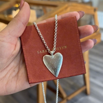 Heart-shaped silver necklace on a red card held by a hand, with a blurred indoor background.