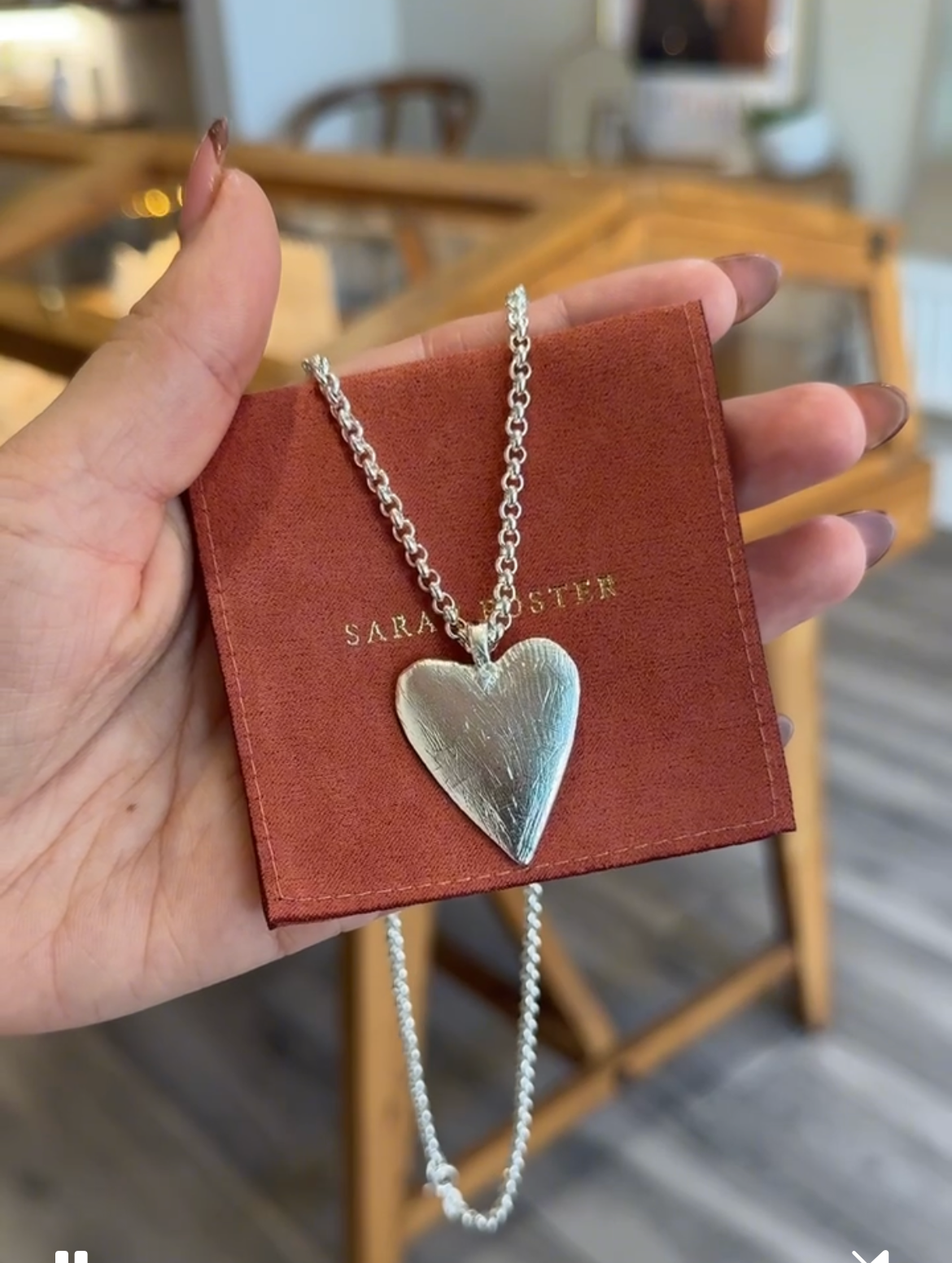 Heart-shaped silver necklace on a red card held by a hand, with a blurred indoor background.