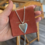 Heart-shaped silver necklace on a red card held by a hand, with a blurred indoor background.